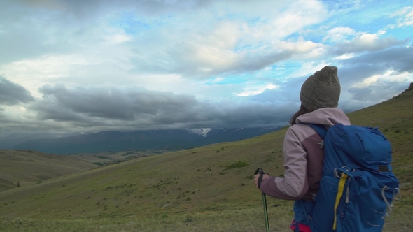 A Girl with a Backpack and Trekking Sticks Stands and Admires the Mountain View