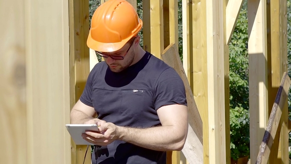 Builder Worker Is Comparing Frame of House with Project in a Flatbed, Looking Up, Wearing Protective alt