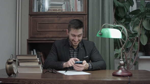 Young Writer at His Desk Happily Messaging on Mobile Phone alt