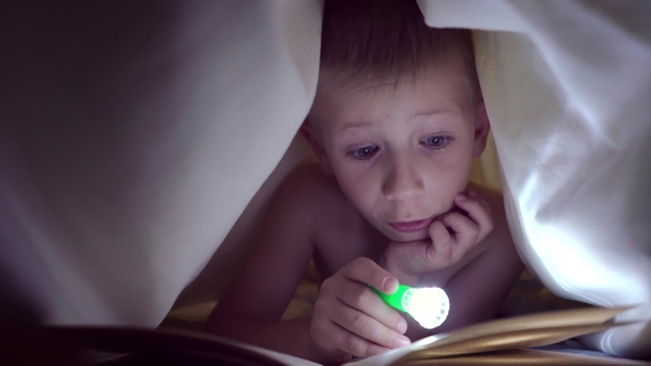 A Child Reads a Book Under Blankets with a Flashlight at Night. Boy with Light Hair and Blue Eyes