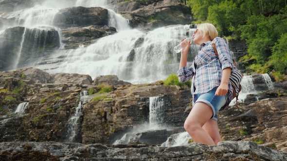 Active Woman Drinks Clean Water Against the Background of the Twindefossen Waterfall in Norway alt