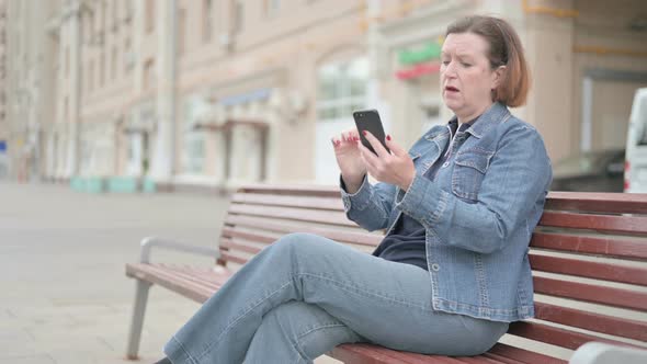 Old Woman Reacting to Loss on Smartphone While Sitting Outdoor on Bench alt