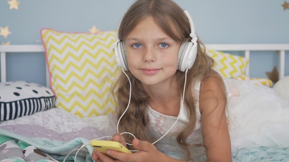 Happy Smiling Girl Lying with Smartphone and Headphones in Bed Listening To Music at Home
