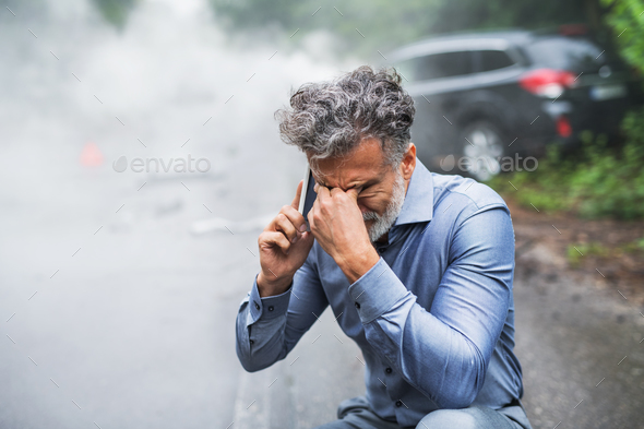 Mature man making a phone call after a car accident, smoke in the background. Stock Photo by halfpoint
