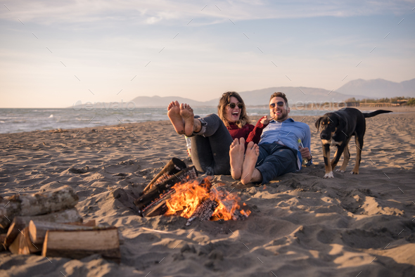 Young Couple Sitting On The Beach beside Campfire drinking beer Stock Photo by dotshock