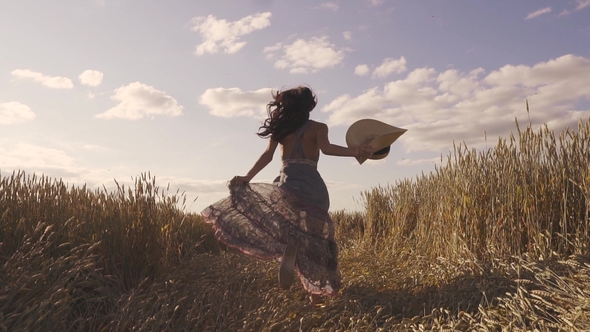 Beautiful Young Woman Running Across the Field with a Straw Hat Turns Around Smiling at the Camera