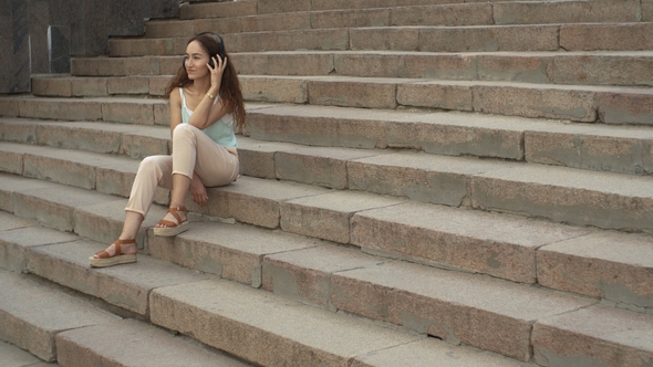 Attractive Brunette Girl Listens Music and Sitting on the Stairs Outside alt
