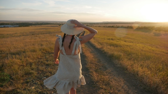 Girl in the Hat and Dress Is on the Field at Sunset alt