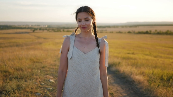 Young Woman in a Hat Stands on the Field During the Sunset alt