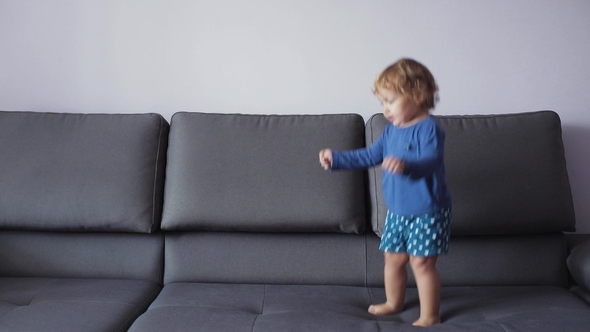 Little Girl with Curly Blond Hair Jumps on Sofa. Blue Clothes. Feels Happy