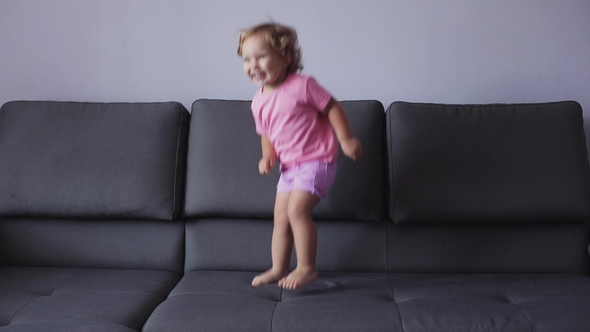 Little Girl with Curly Blond Hair Jumps on Sofa. Blue Clothes. Feels Happy