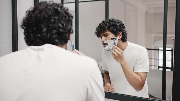 Young Indian Man Shaves Beard with Razor Looking in Mirror alt