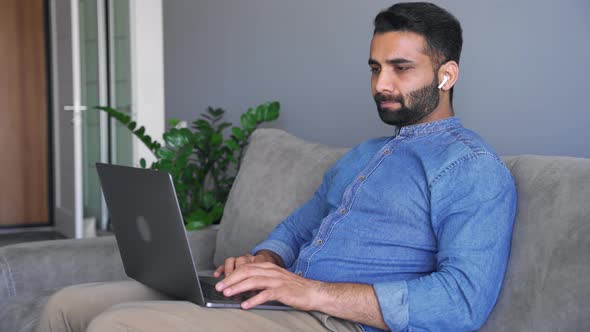 Young Adult Indian Man Typing on Laptop Keyboard Sitting on Sofa Couch alt