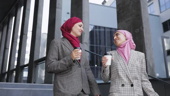Two Young Muslim Women Wearing Hijab Headscarf Look at Phone, Walking Together Down Stairs Drinking