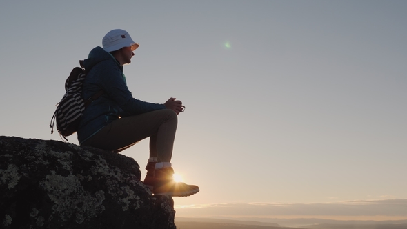 A Traveler Drinks Tea From a Mug at the Peak of the Mountain. Sits on a Rock, in the Background alt