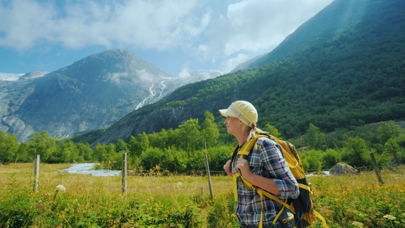 An Active Woman with a Backpack Walks Against the Backdrop of the Mountains and the Briksdal Glacier alt