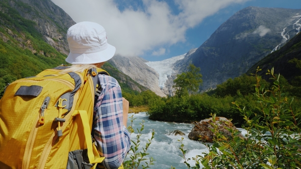 A Tourist with a Yellow Backpack Looks at a Beautiful Glacier at the Top of the Mountain alt