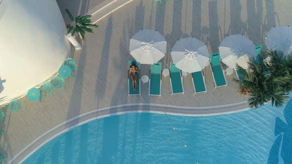 Aerial: Beautiful Girl Lies on a Chaise-longue By the Pool, Drinking a Cocktail