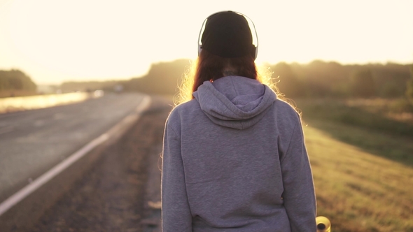 Portrait of a Red-haired Girl in Headphones Walking Along the Roadside at Sunset. Back View