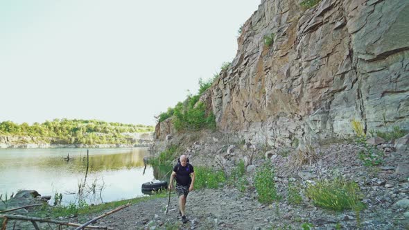 adult tourist is walking along the bank of the lake with a stick in his hand and with a backpack alt