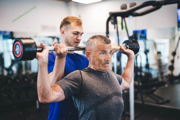 Senior man lifting weights with help of gym assistant. Stock Photo by photocreo