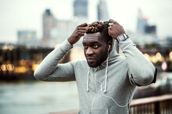 Black man runner with earphones on the bridge in a city, putting hood on head. Stock Photo by halfpoint
