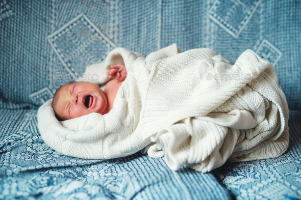 Crying newborn baby lying on a sofa, covered by a white blanket. Side view. Stock Photo by halfpoint