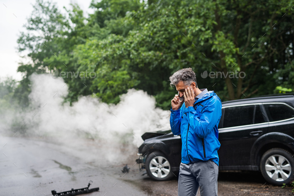 Mature man making a phone call after a car accident, smoke in the background. Stock Photo by halfpoint