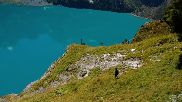 Arial shot of a blonde woman with backpack hiking down a lush mountain at a blue mountain lake in th alt