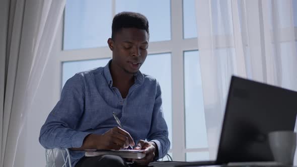 Focused African American Employee Writing Ideas in Sketchpad Listening to Video Chat on Laptop alt