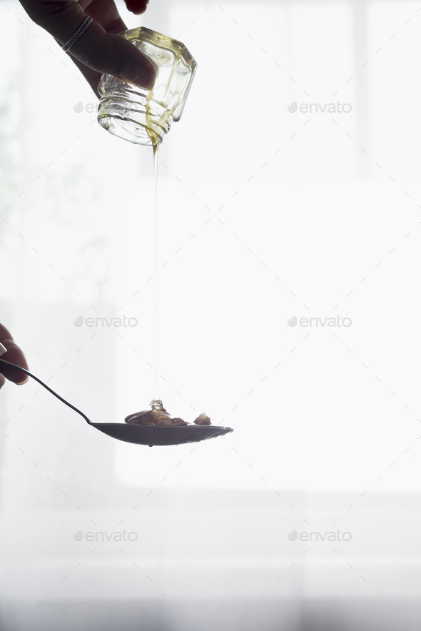 In the hands of the glass jar with honey pours out into a spoon over light window - Stock Photo - Images