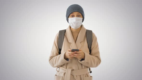 Woman Dressed in a Coat Wearing Medical Mask Using Phone and Walking on Gradient Background. alt