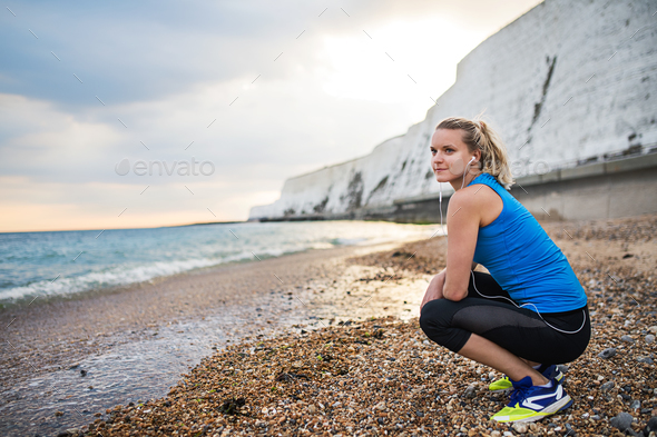 Young sporty woman runner with earphones resting on the beach outside. Stock Photo by halfpoint
