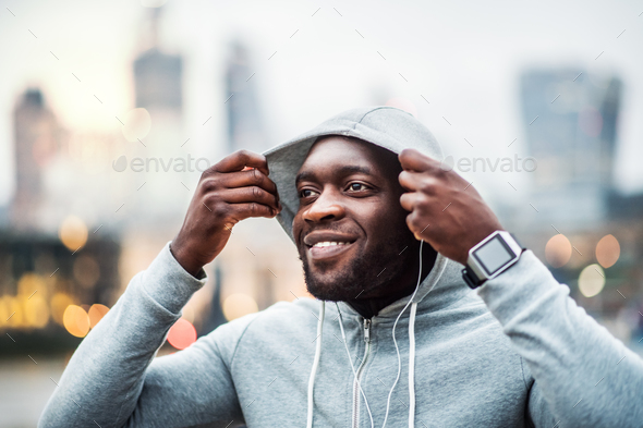 Black man runner with earphones on the bridge in a city, putting hood on head.