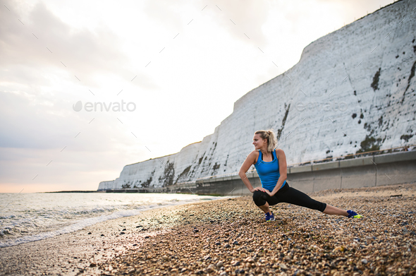 Young sporty woman runner in blue sportswear stretching on the beach outside. Stock Photo by halfpoint