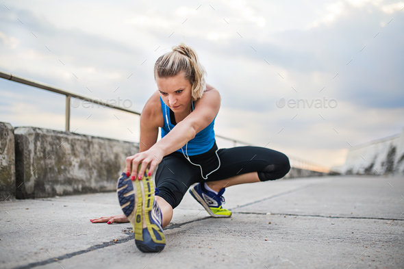 Young sporty woman runner with earphones stretching outside. Stock Photo by halfpoint