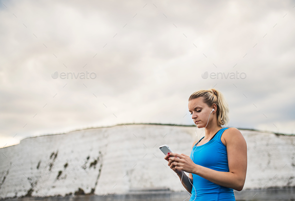 Young sporty woman runner with earphones and smartphone standing on the beach. Stock Photo by halfpoint