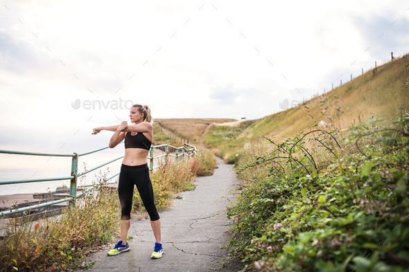Young sporty woman runner in black activewear standing outside in nature, stretching. Stock Photo by halfpoint