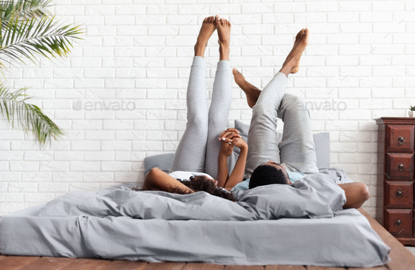 Happy couple lying on bed at home raising legs up Stock Photo by Prostock-studio
