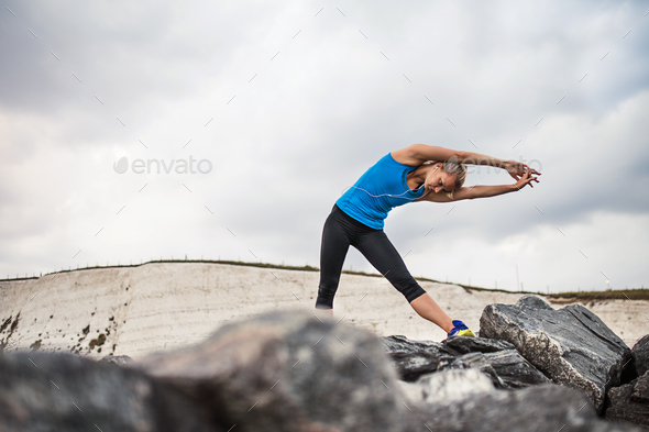 Young sporty woman runner with earphones standing on the rocks outside, stretching. - Stock Photo - Images