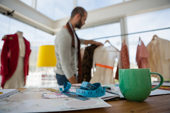 Tape measure by cup and designs on table with designer working in background Tape measure by cup and designs on table with designer working in background