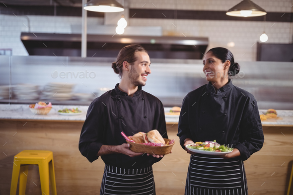 Smiling young wait staff looking at each other while holding fresh food in coffee shop