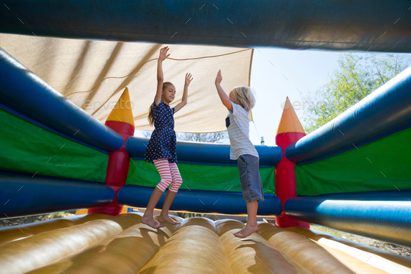 Cheerful siblings with arms raised jumping on bouncy castle