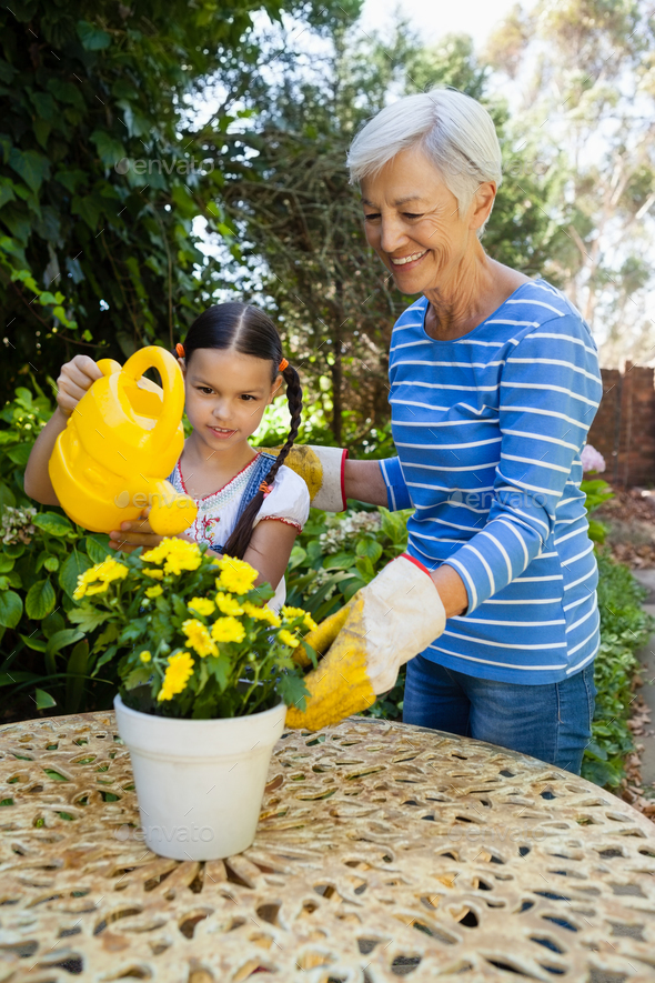 Smiling senior woman standing by granddaughter watering yellow flowers on table Smiling senior woman standing by granddaughter watering yellow flowers on table