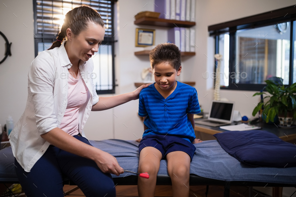Young female therapist examining boy with reflex hammer on knee while sitting on bed