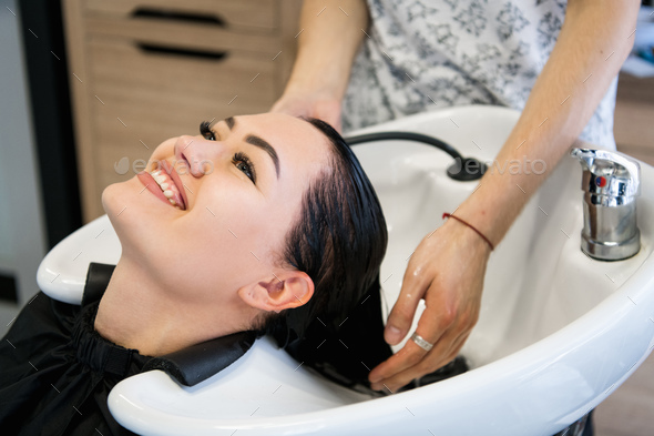 Young brunette girl smiling and enjoying hair washing in hairdressing salon.  Stock Photo by romankosolapov