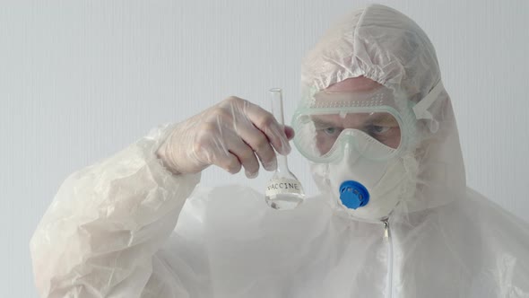 Virologist in Protective Mask and Costume Looking Vaccine Flask in Hands. Biochemist Watching alt
