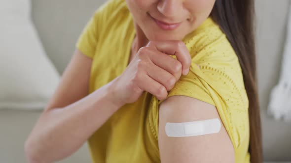 Happy asian woman sitting on sofa showing arm with plaster after vaccination alt