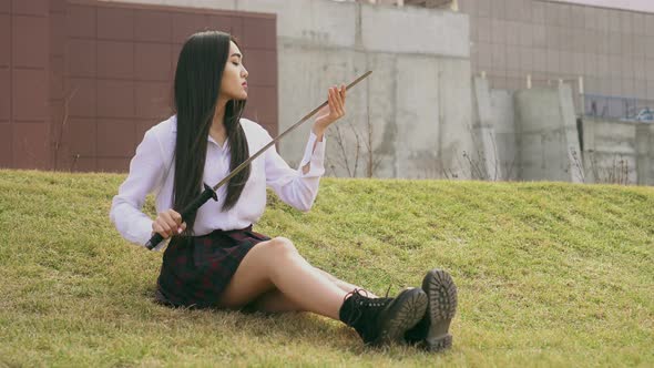 Chinese People Is Sitting on Grass and Strokes Blade of a Sword in Short Unifom alt