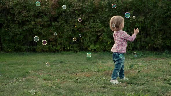 Little Baby Girl Plays with Soap Bubbles and Laughs Outdoors in Summer Park